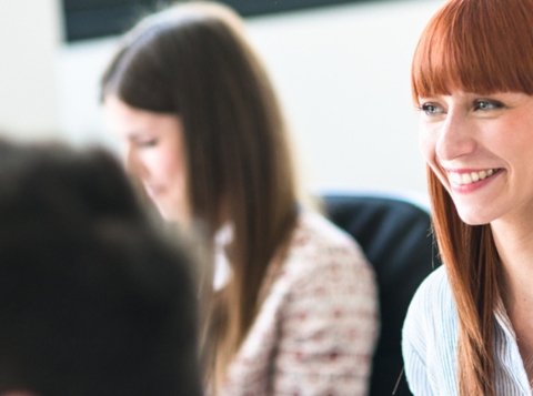 Red headed woman sat at a desk smiling with colleagues around her