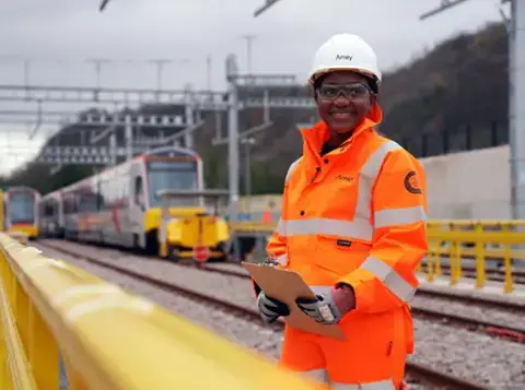 Amey worker in hi-vis PPE holding a clipboard and smiling at a rail depot with a train in the background