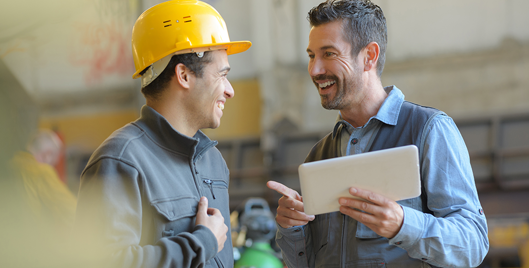 Smiling man holding a tablet talking to another smiling man wearing a hard hat in a warehouse