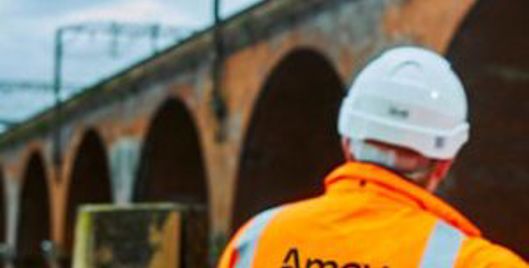 Amey worker in orange high-visibility jacket and white hard hat surveying a brick railway viaduct