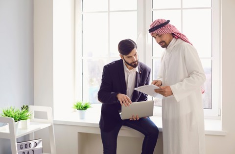 Two men in an office discussing documents on a laptop