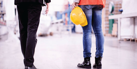 Person in suit walking next to person in high vis holding hard hat in a warehouse
