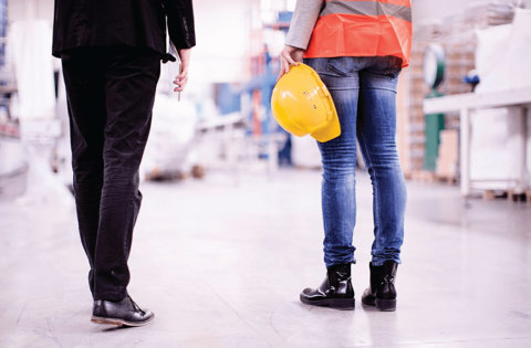 Person in suit walking next to person in high vis holding hard hat in a warehouse