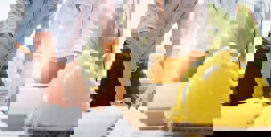 Two people working on health and safety documents with a hard hat on the table next to them