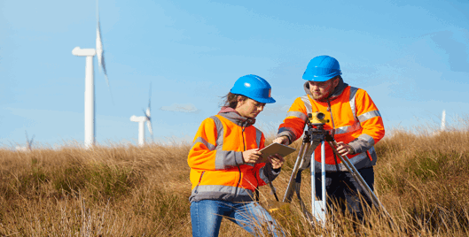 Two people wearing orange high vis jackets looking at a clipboard in a wind farm field